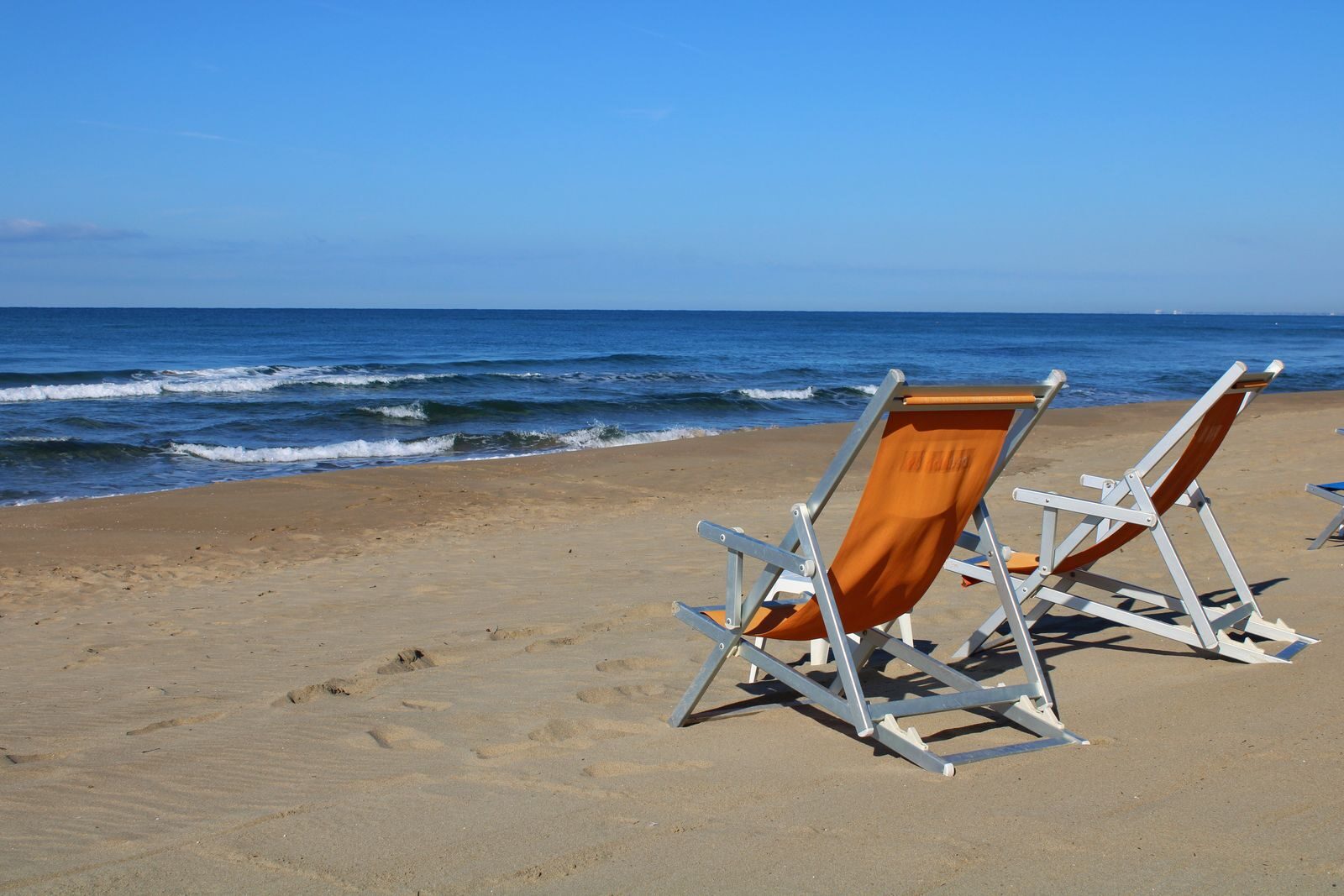 Two empty deck chairs on a sandy beach under clear blue skies in Latina, Italy.